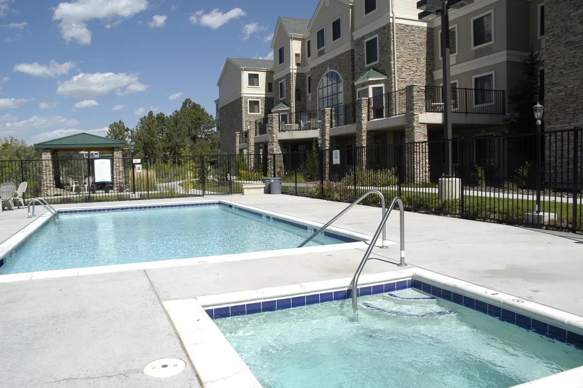 This image features an inviting exterior view of the swimming pool and adjacent hot tub, surrounded by lush greenery and the apartment building in the background. Guests can be seen swimming, sunbathing, and relaxing in the hot tub, conveying both leisure and social interaction.