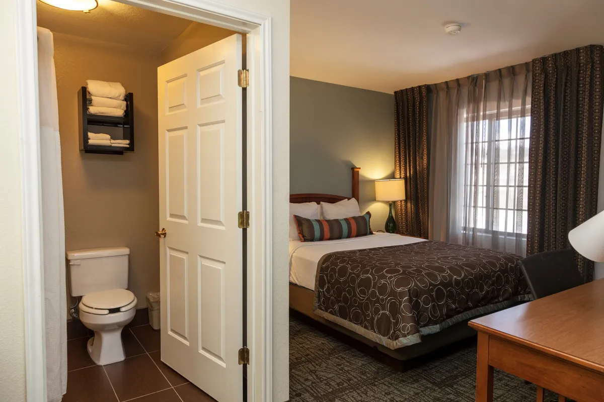 This image centers on a cozy bedroom scene featuring a king bed adorned with decorative pillows. A soft glow from the bedside lamp adds warmth, while a desk provides a workspace next to a window with curtains. The bathroom door is visible, adding to the sense of privacy and convenience.
