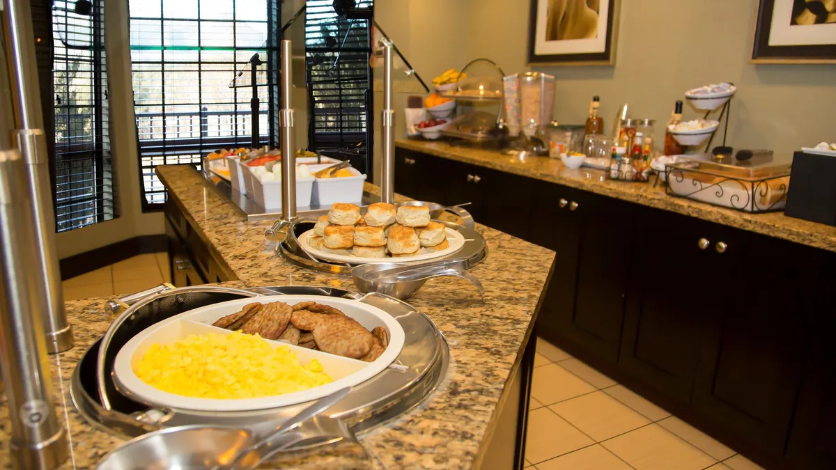 A welcoming interior scene featuring a breakfast buffet spread on a granite countertop. The area showcases an array of baked goods and breakfast items, with guests enjoying a relaxed morning atmosphere, hinting at social interactions and the beginning of a lively day.